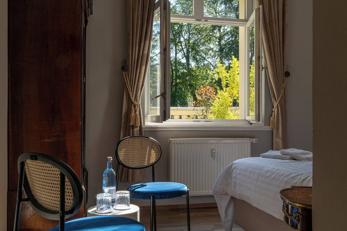 Une chambre à coucher avec un lit, une fenêtre avec des rideaux, une chaise, une petite table avec une bouteille et des verres, et un radiateur.