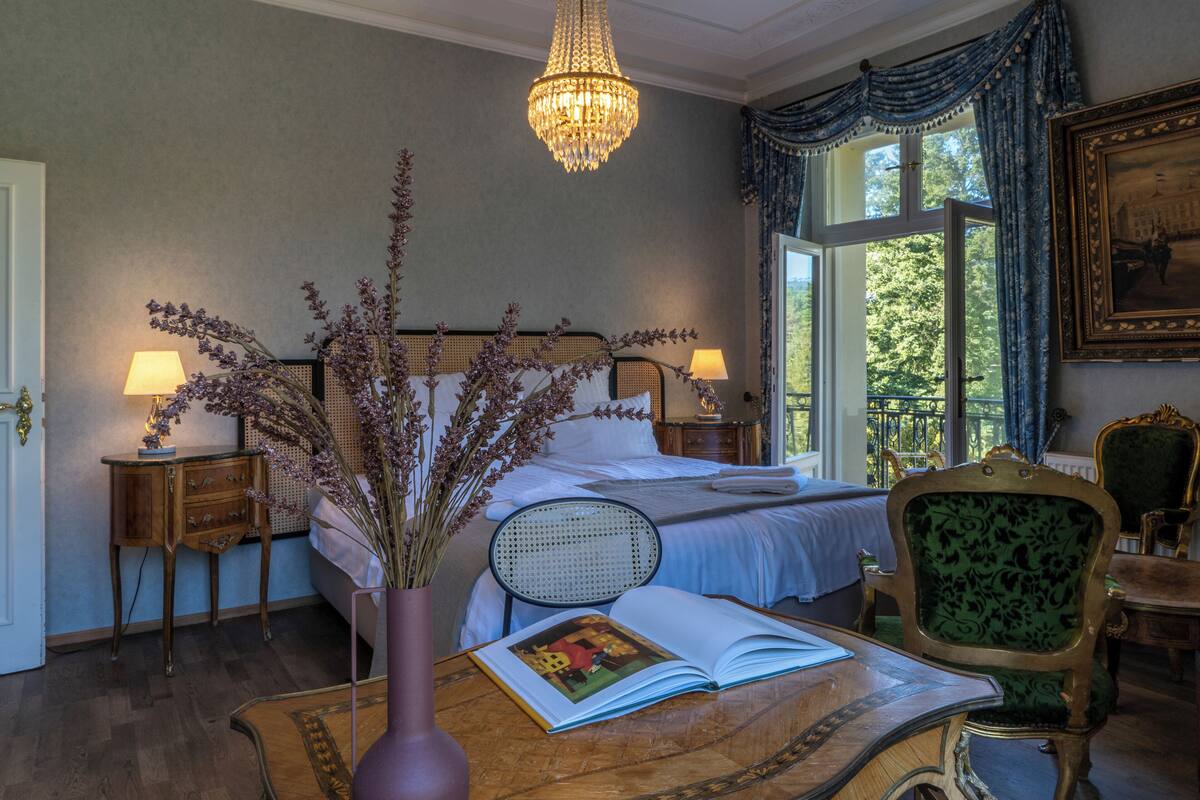 Une chambre à coucher avec un lit, des lampes de chevet, un vase avec des fleurs, une chaise et une table sur laquelle se trouve un livre.