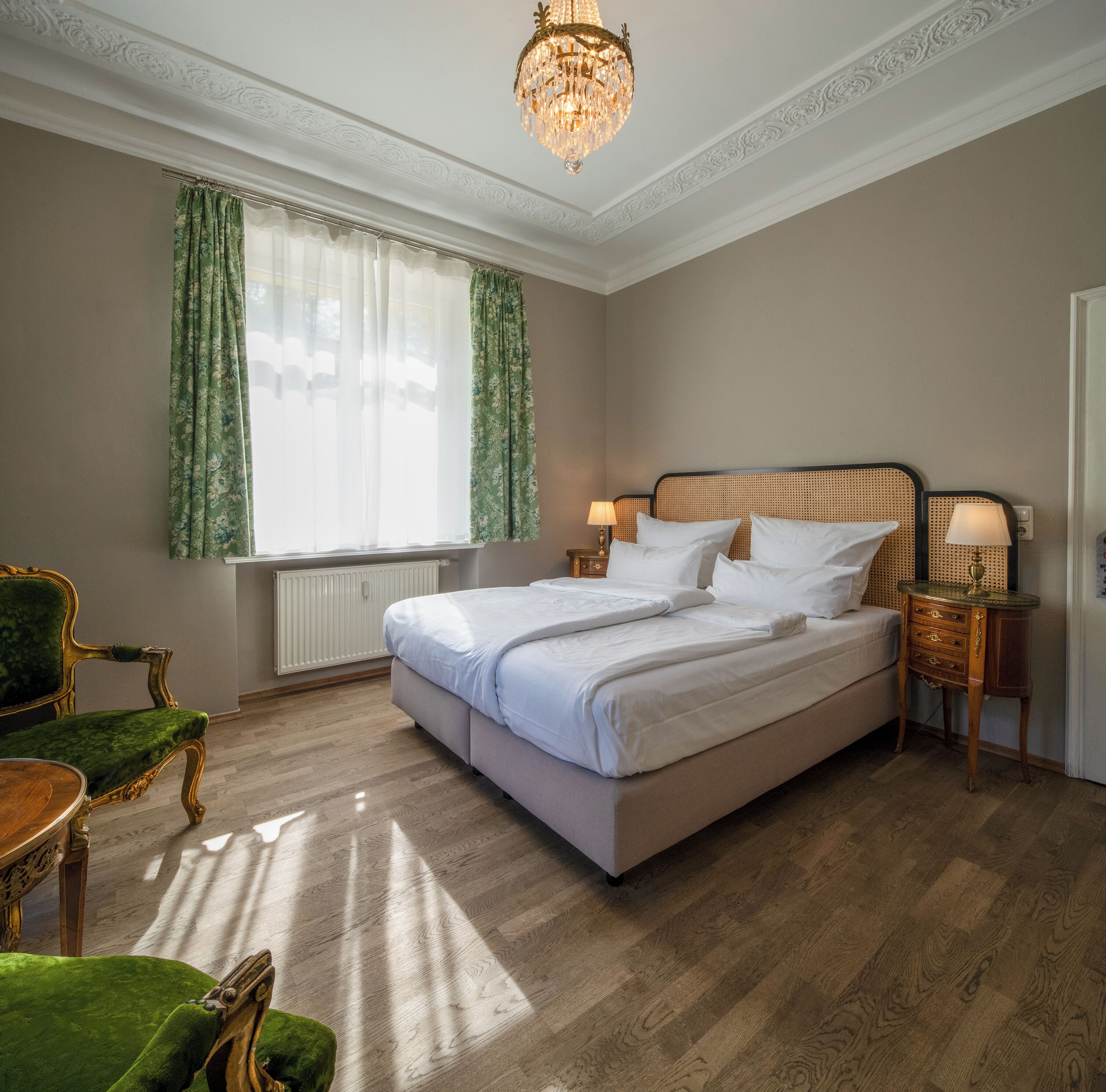 A neatly arranged bedroom with a large bed, two bedside tables with lamps, a green upholstered chair, and a chandelier.