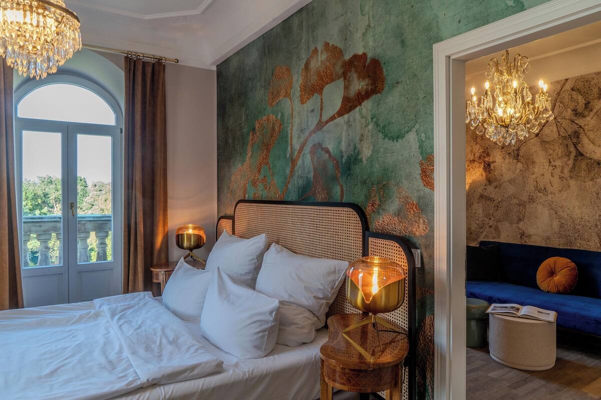 Une chambre à coucher avec un lit, une table de chevet, une fresque murale, un lustre et une vue par la fenêtre.