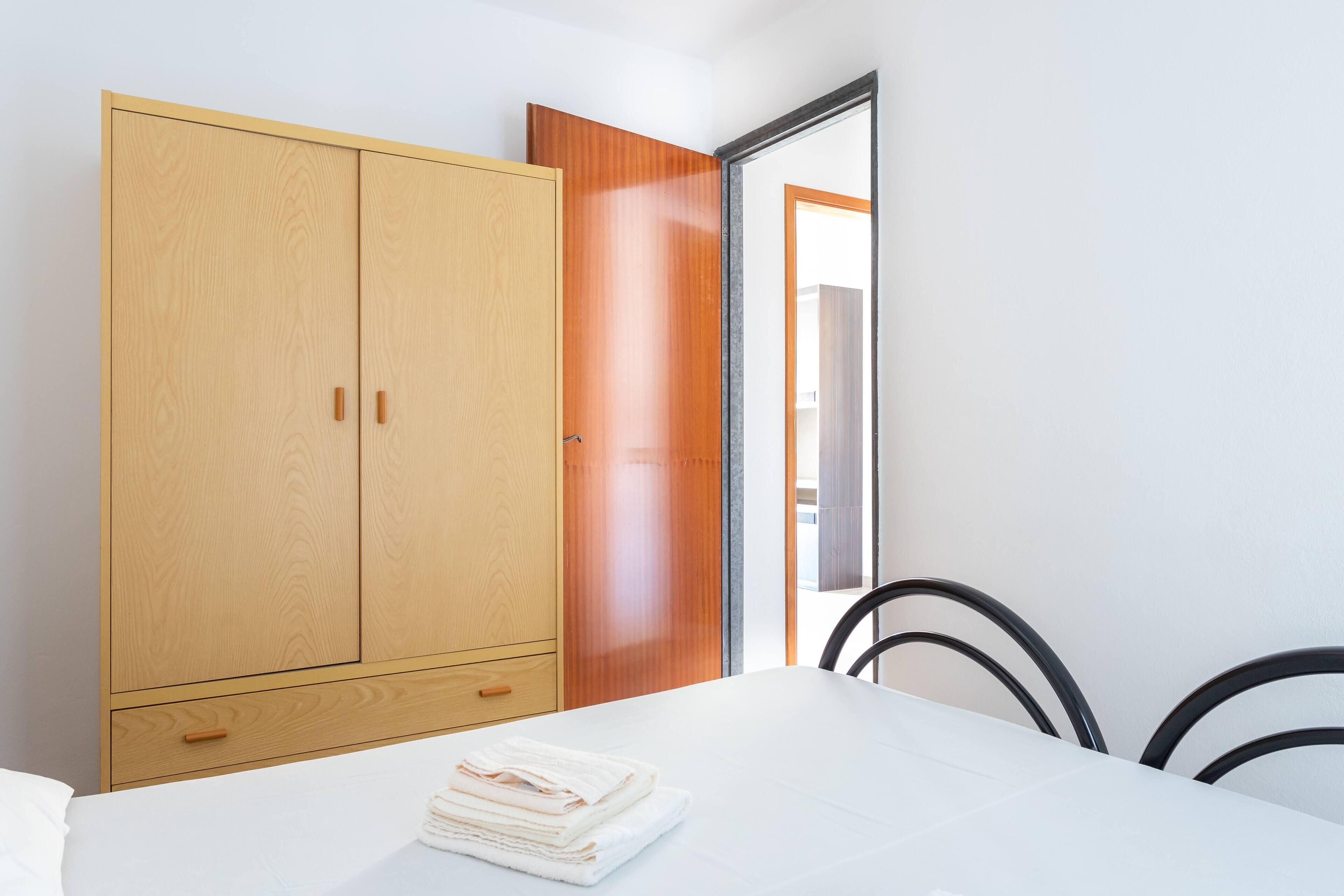 Une chambre avec une grande armoire en bois, un lit recouvert de draps blancs et une vue sur l’extérieur par une porte ouverte.