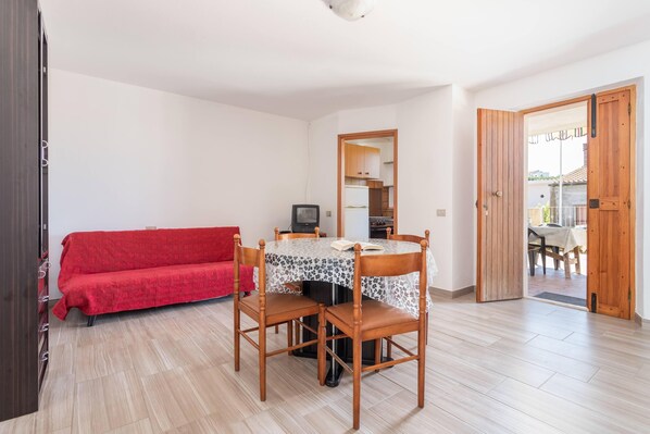 A room with a dining table, chairs, a red sofa, a television, and a kitchen area visible through an open door.