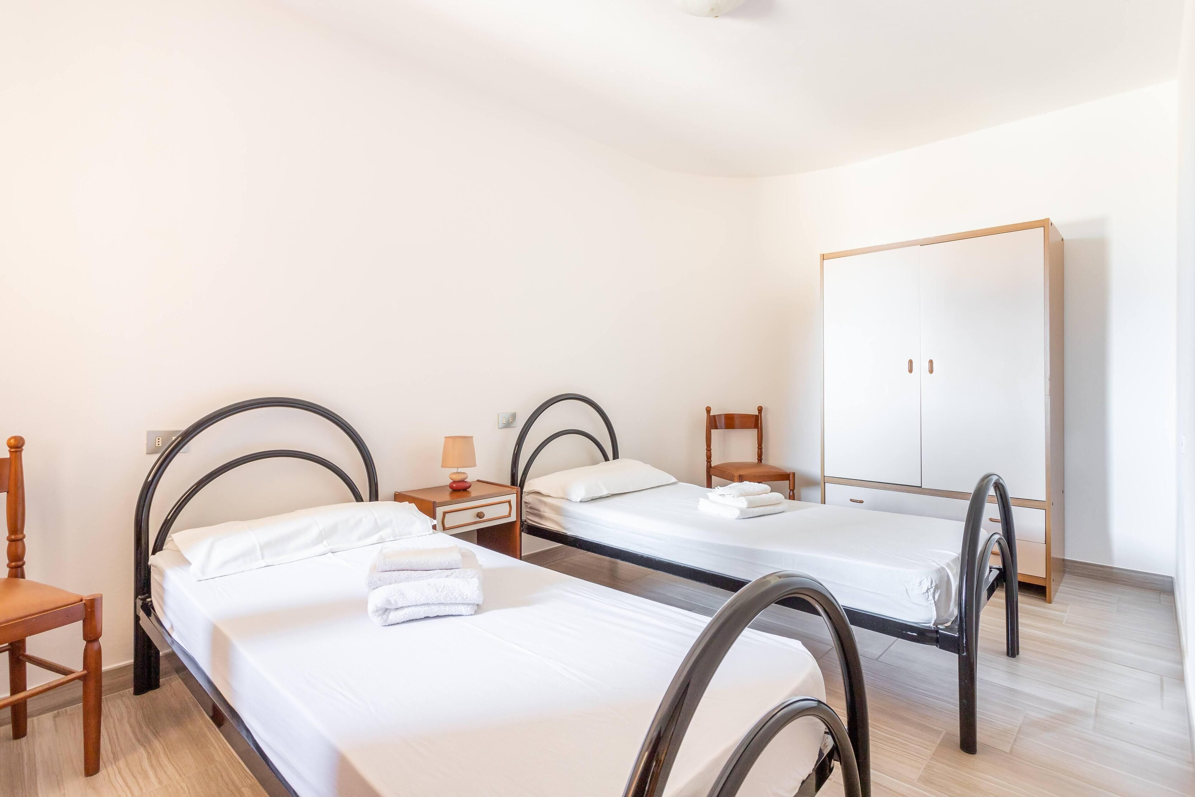A hotel room with two single beds, a wooden chair, a wardrobe, and a nightstand with a lamp.