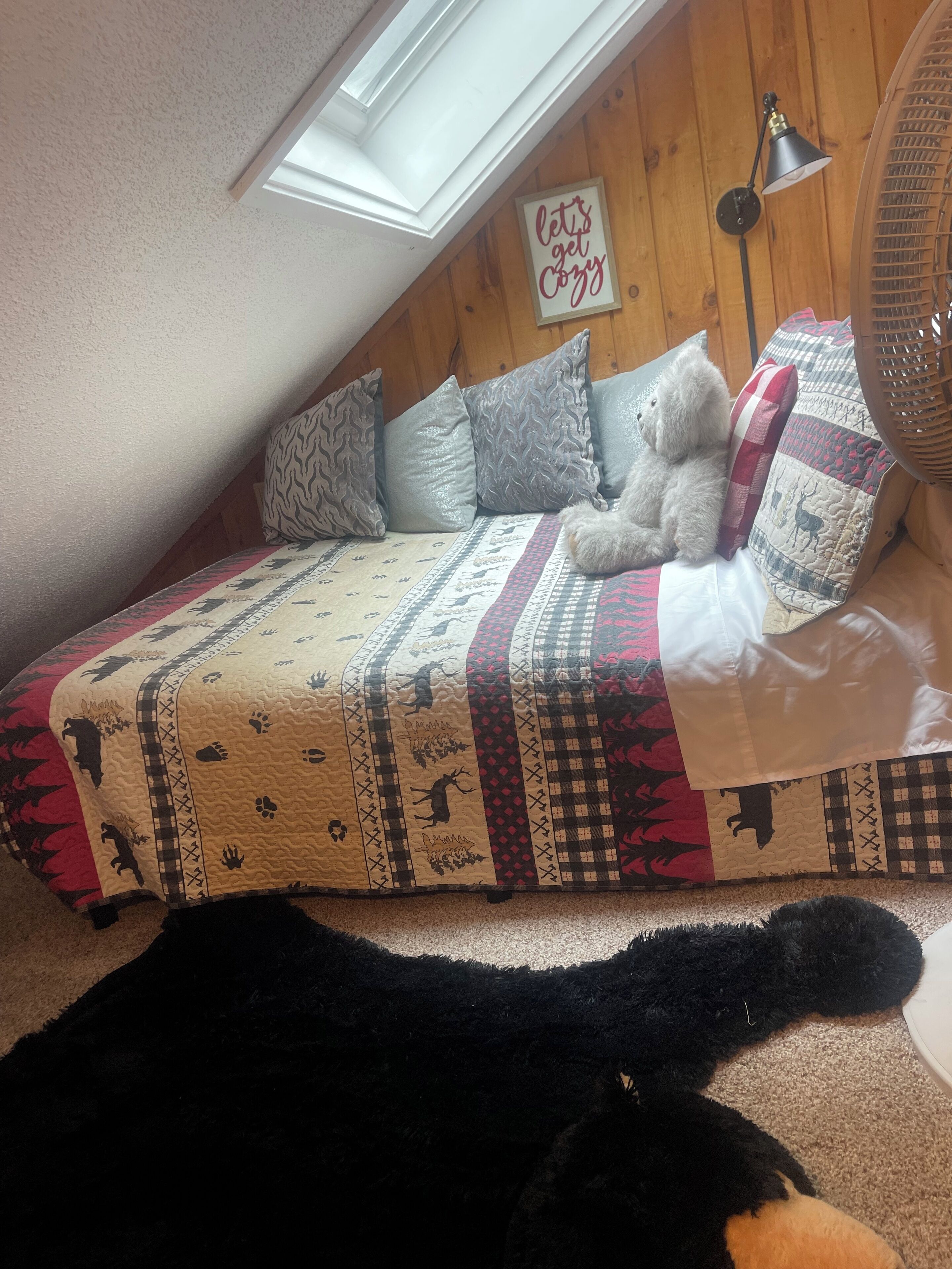 Une chambre à coucher chaleureuse aménagée en mezzanine, avec un plafond en pente, un grand lit recouvert d’une literie à motifs, un ours en peluche et une pancarte sur laquelle on peut lire : « Installons-nous confortablement ».