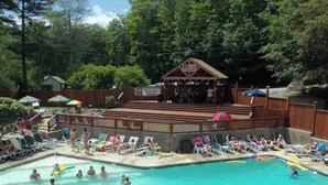 A swimming pool area with multiple pools, lounge chairs, and a covered stage with an American flag. - White Mountains Chic Chateau: Updated 3BR+Loft, Sauna, Fireplace & Resort Perks (Campton)