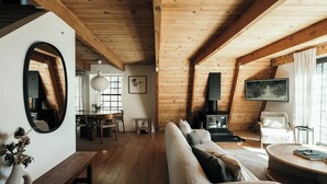 Ein gemütliches Blockhaussinnere mit Holzbalkendecke, einer Couch, einem runden Tisch, einem Kamin und einem großen Spiegel.