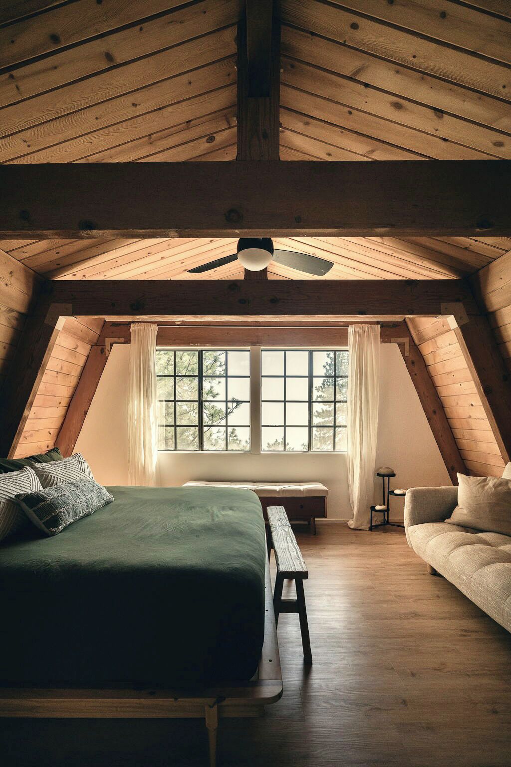 Un dormitorio tipo loft con techo inclinado, una cama, un banco, un sofá y una ventana con cortinas.