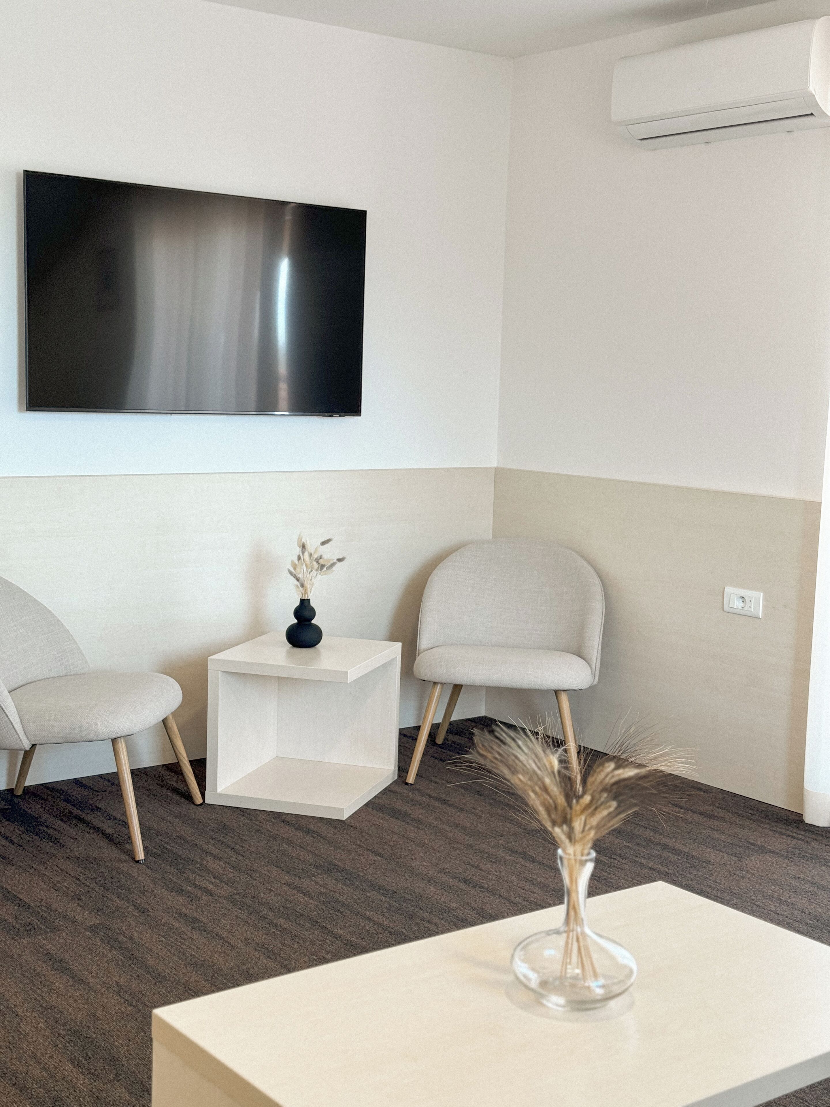 A modern hotel room with a flat-screen TV mounted on the wall, two upholstered chairs, a small side table with a vase, and a wooden floor.