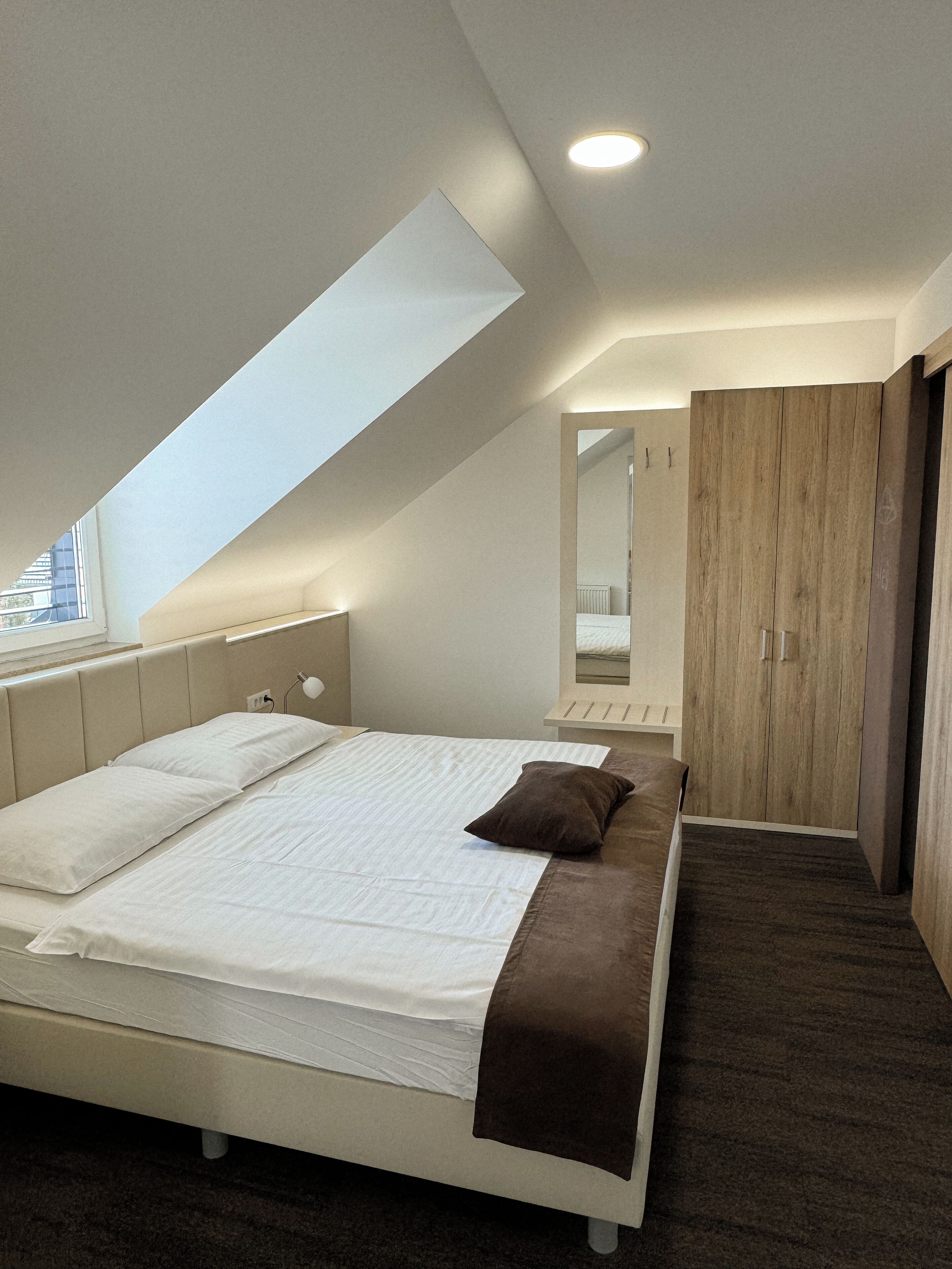 Un dormitorio moderno con techo inclinado, una cama grande con ropa blanca, un plaid marrón, un armario de madera y un tragaluz.