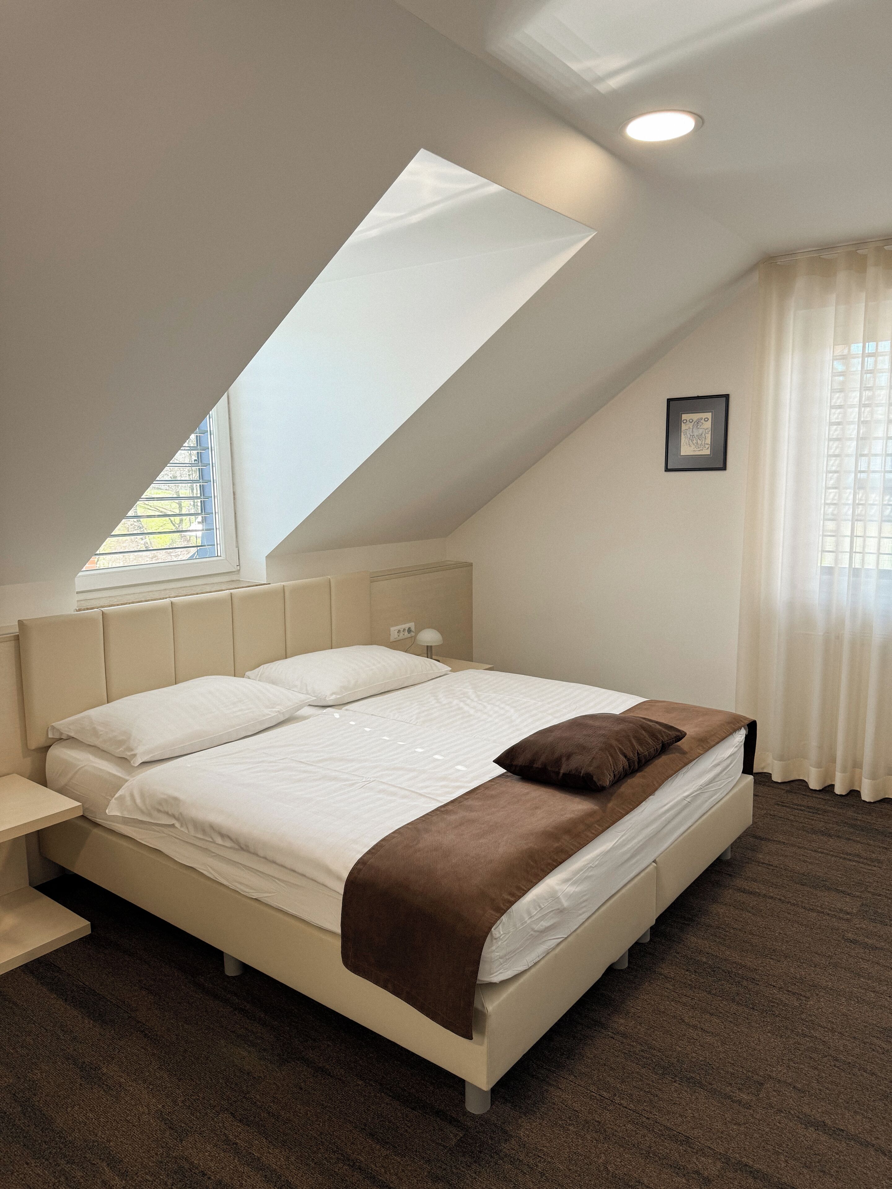Un dormitorio con techo inclinado, una cama con ropa de cama blanca y un plaid marrón, una lámpara de noche y una ventana con persianas.