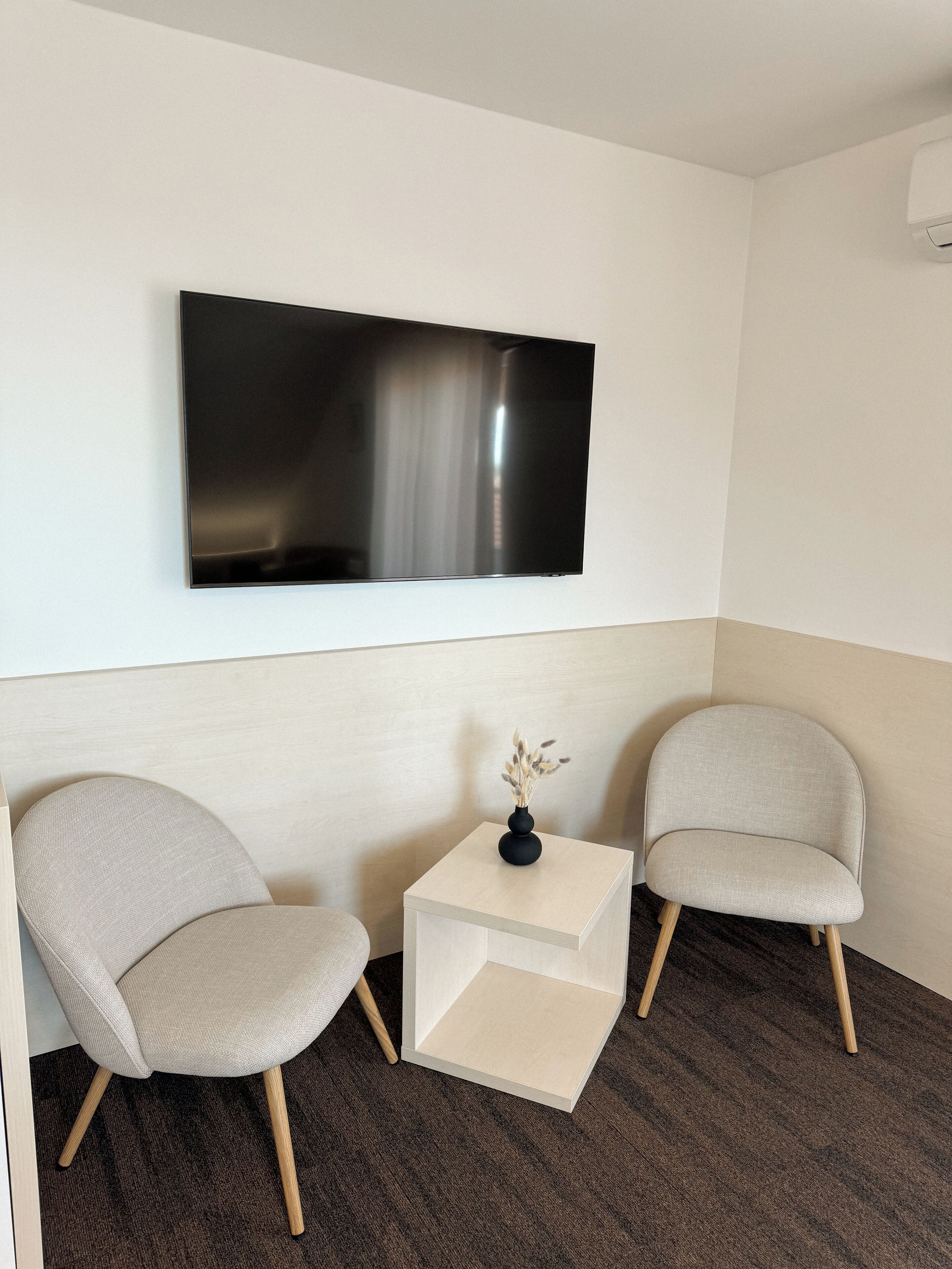 A modern room with a flat-screen TV mounted on the wall, two beige armchairs, a small wooden side table with a vase, and a dark brown carpet.