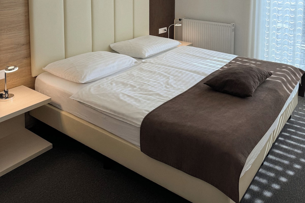 Moderni hotellihuone, jossa on suuri sänky, yöpöytä, seinään kiinnitetty valaisin ja ikkuna, jossa on verhot.