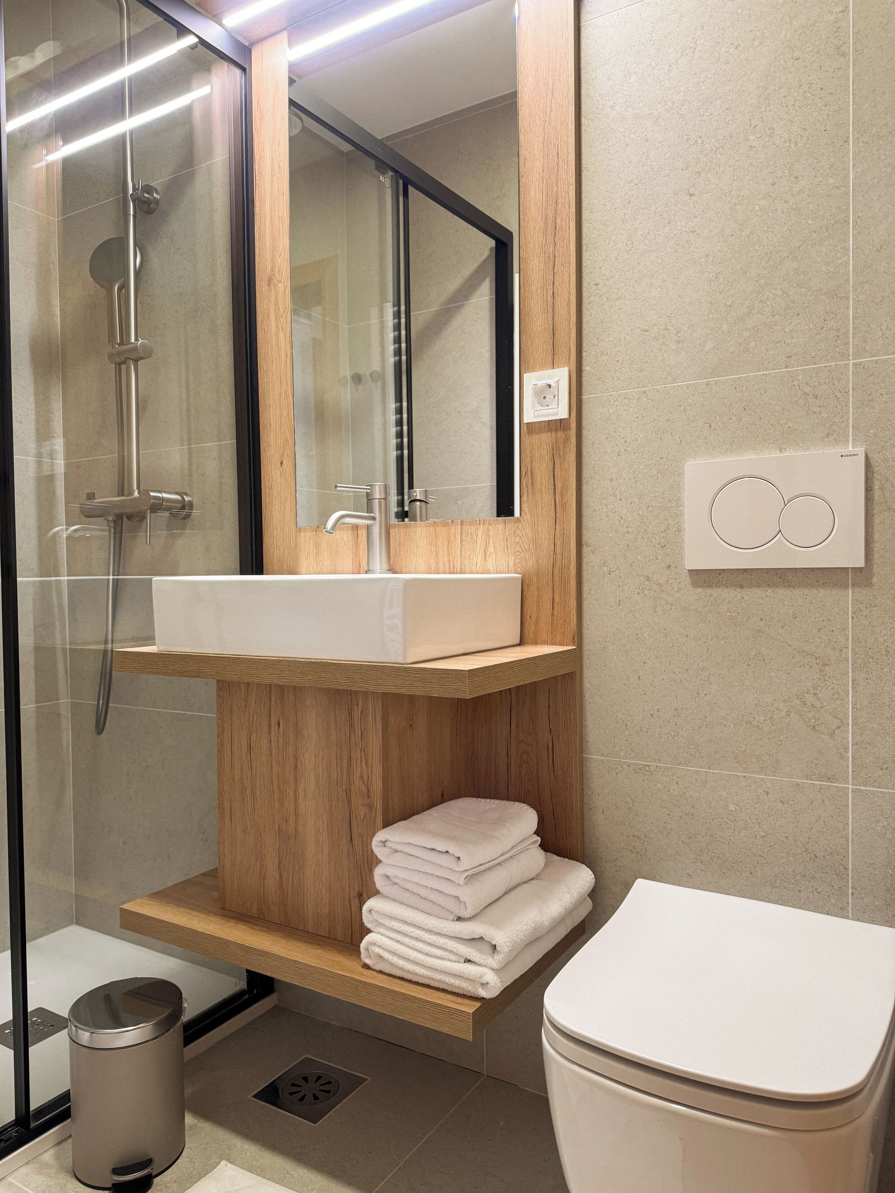 Un baño moderno con ducha de vidrio, tocador de madera con lavamanos, inodoro y espejo.