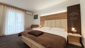 Habitación de hotel con una cama grande, cabecera de madera, mesita de noche con lámpara, una silla y una ventana con cortinas.