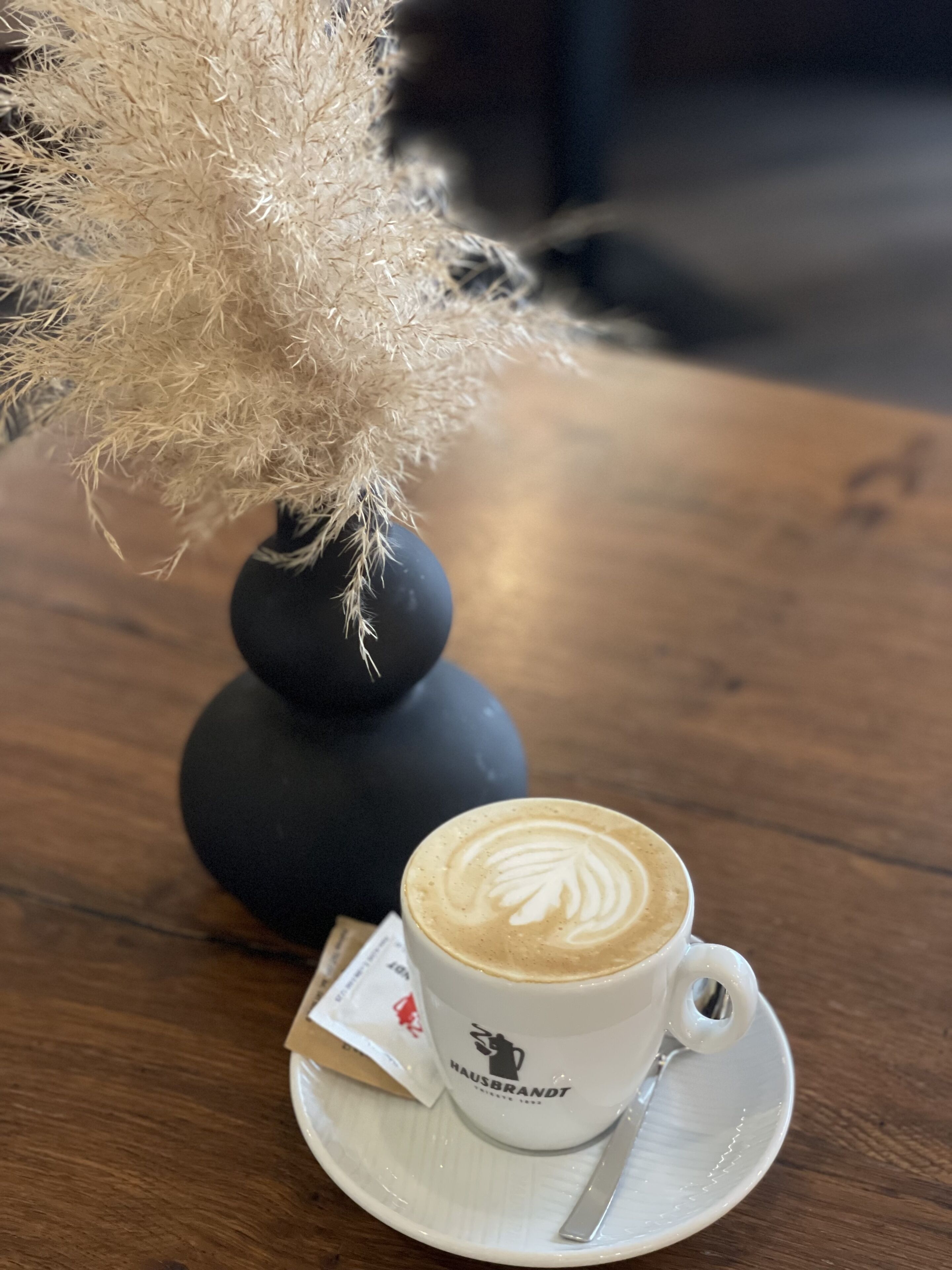 A cup of coffee with a leaf design on the foam, a saucer, and a spoon on a wooden table.