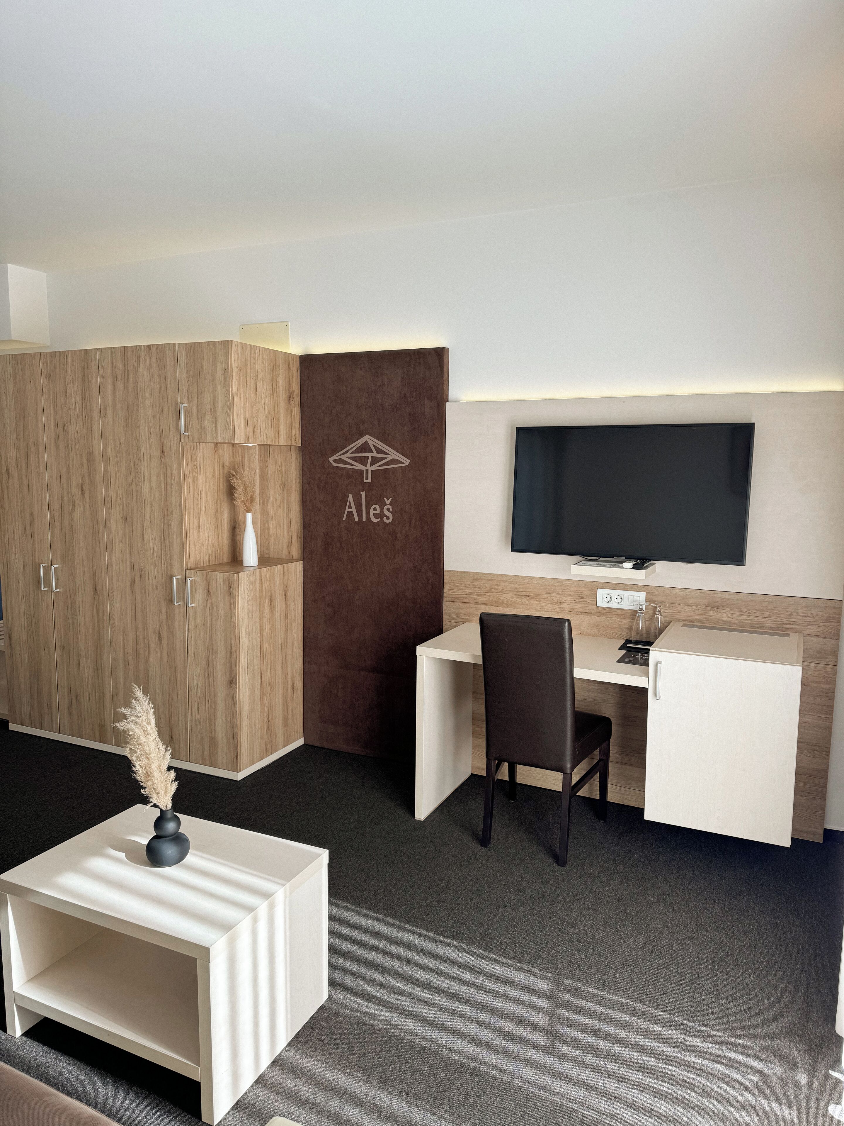 Une chambre d’hôtel moderne équipée d’une armoire en bois, d’un bureau avec une chaise, d’un téléviseur à écran plat et d’une petite table d’appoint agrémentée d’un vase.