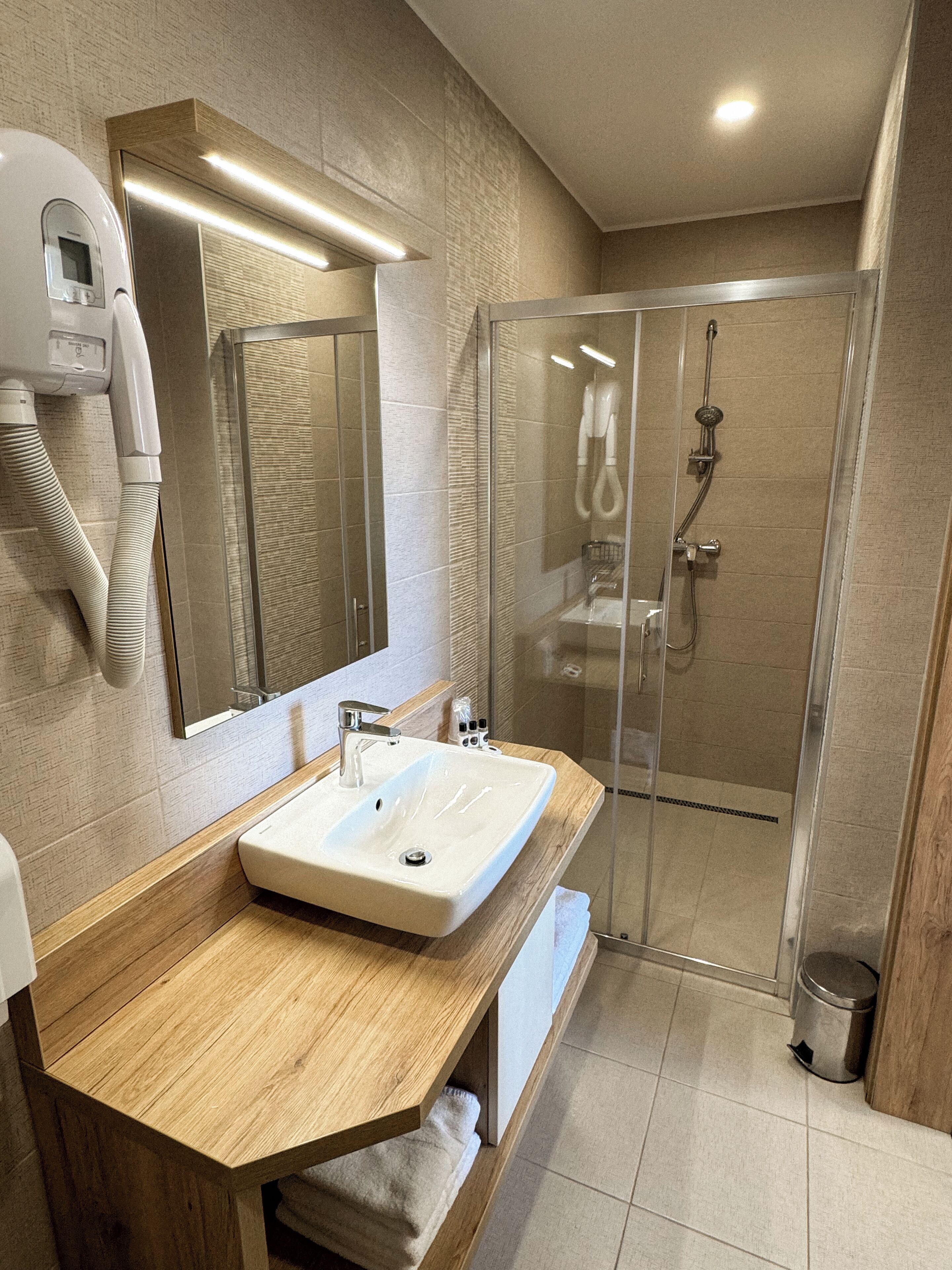 A modern bathroom with a wooden vanity, a large mirror, a built-in shower, and a hairdryer mounted on the wall.