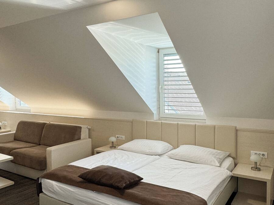 A modern bedroom with a sloped ceiling, a bed with white bedding, a brown sofa, and a small bedside table with a lamp.