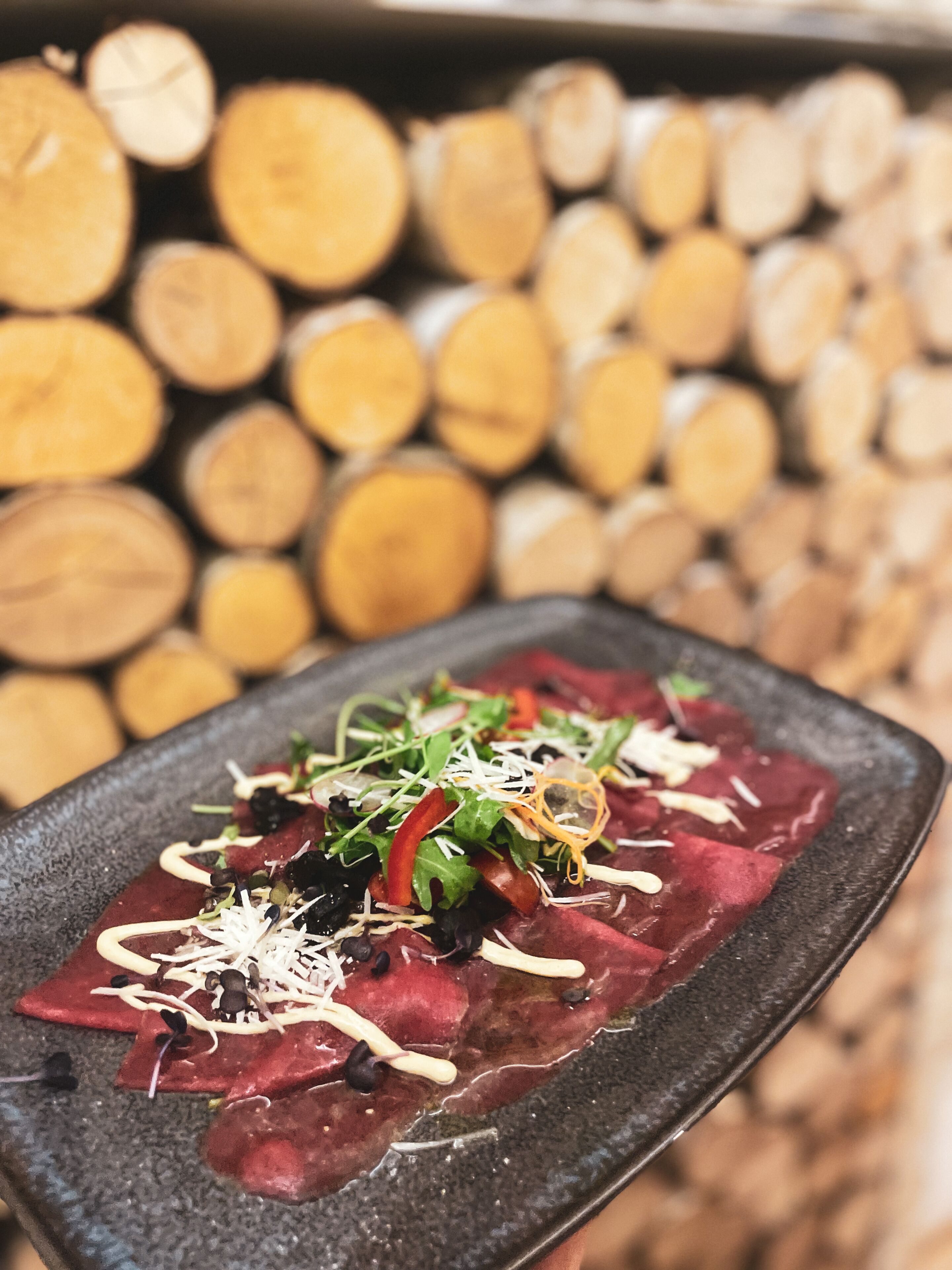 Un plato rectangular con carne roja en lonjas finas, decorado con vegetales verdes, pimientos rojos y queso, sobre un fondo de leña apilada.