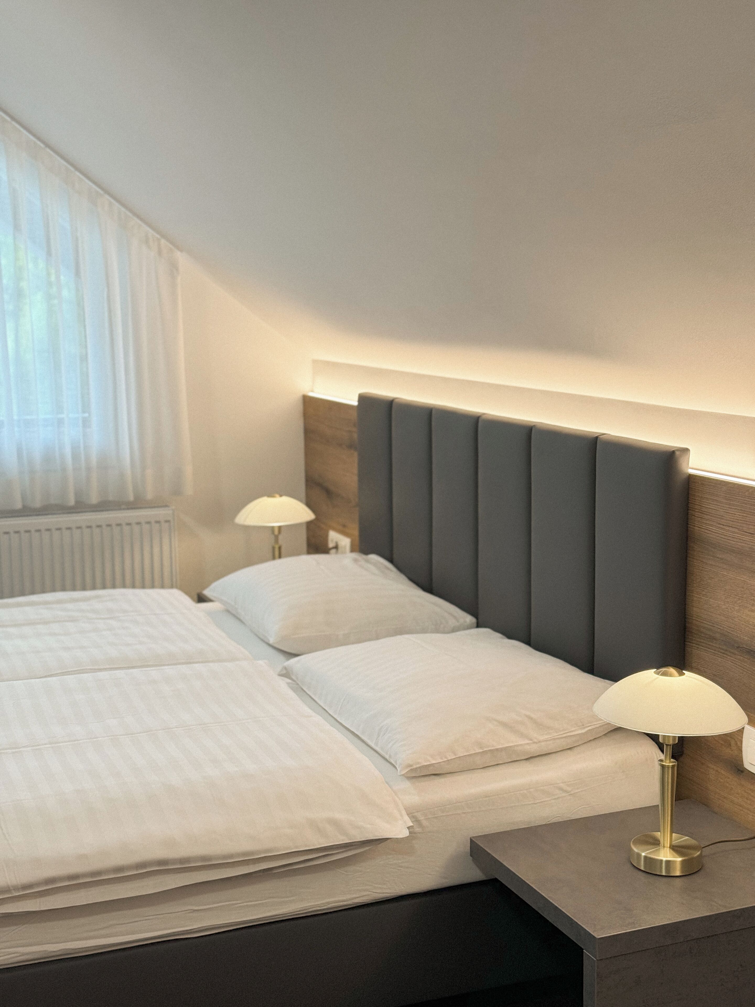 Moderni hotellihuone, jossa on suuri sänky, kaksi yöpöytävalaisinta ja ikkuna, jossa on läpikuultavat verhot.