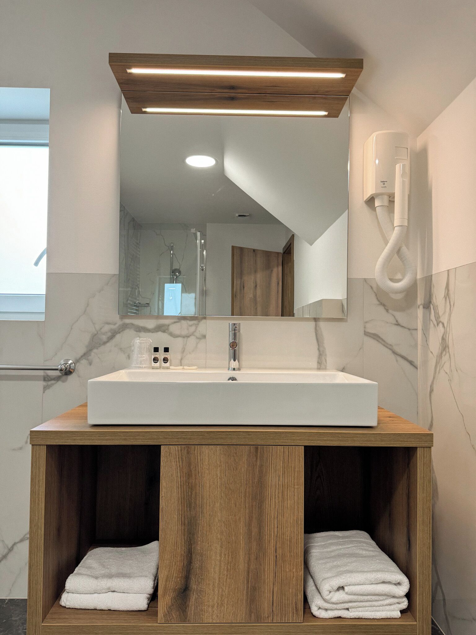 Une salle de bain moderne dotée d’un grand lavabo rectangulaire, d’un meuble-lavabo en bois, de murs en marbre et d’un miroir avec éclairage intégré.