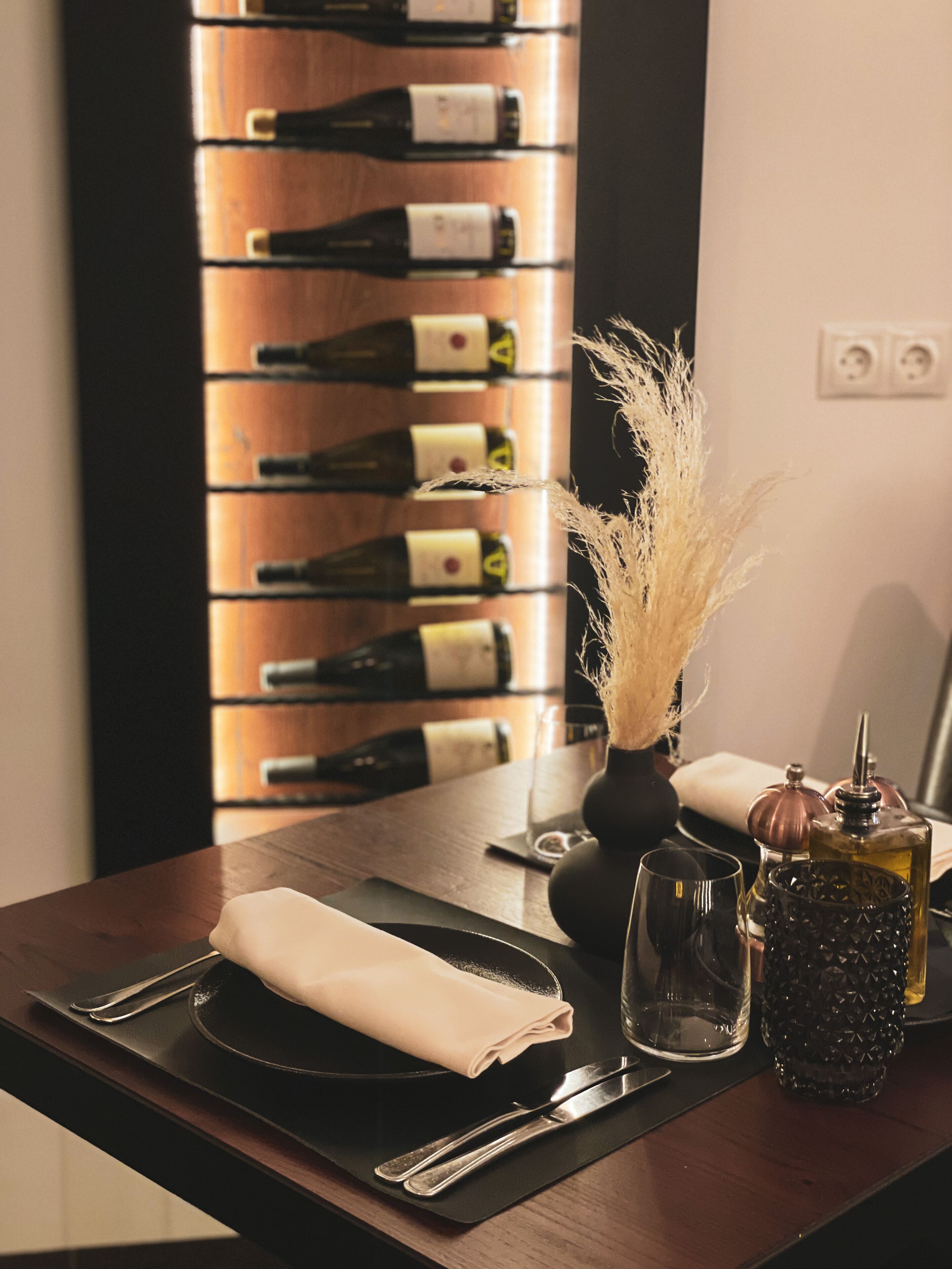 Une table à manger avec une serviette pliée, un verre, une bouteille et un vase contenant des plantes séchées. Au fond, on aperçoit un meuble à vin avec plusieurs bouteilles.