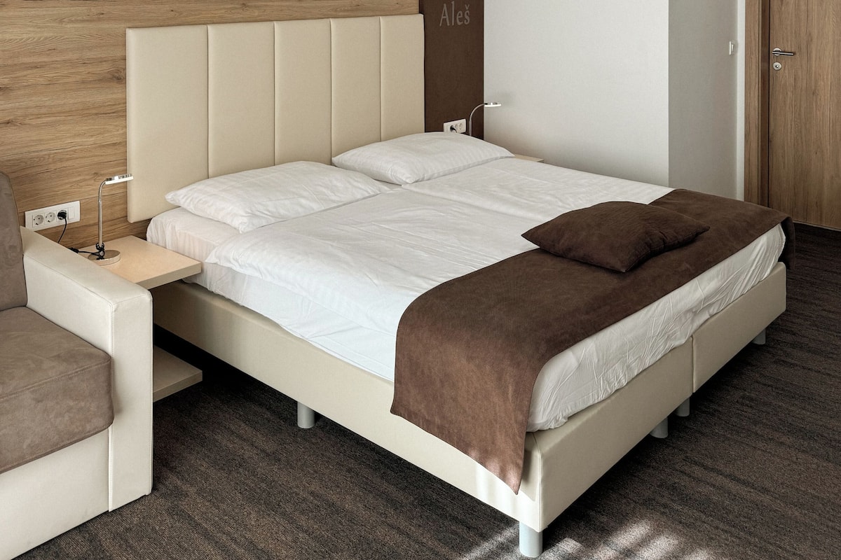 Moderni hotellihuone, jossa on suuri sänky, yöpöytä, seinälle kiinnitetty taideteos ja ovi. Lisäksi huoneessa on sängynpääty, joka on valmistettu puusta.