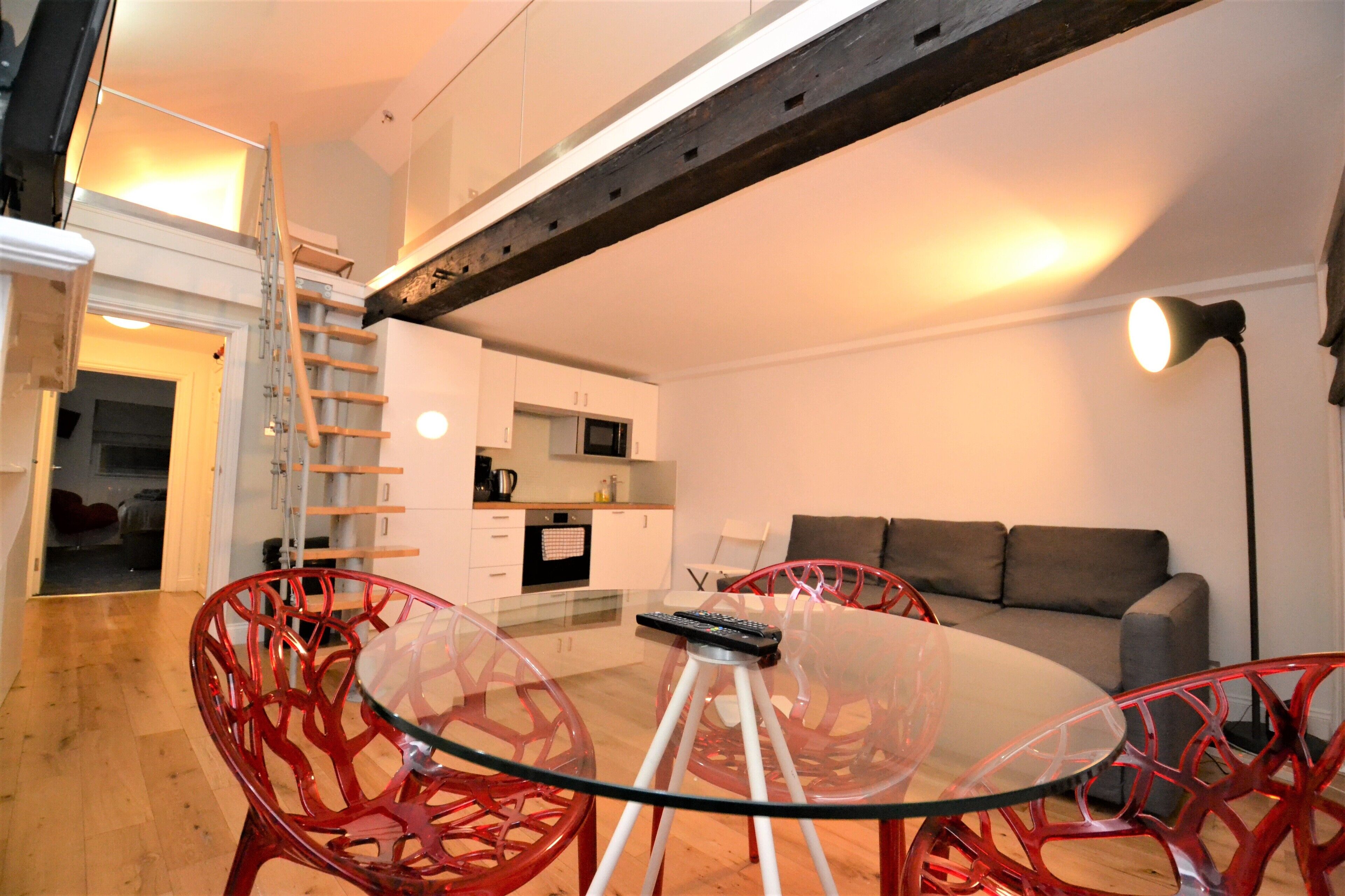 A modern living room with a glass dining table, red chairs, a sofa, and a staircase leading to an upper level.
