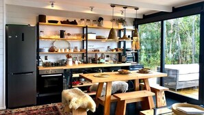 A modern kitchen with wooden shelves, a dining table, and a large window overlooking a forest. - Calma Cabins (Puerto Octay)