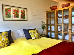 A bed with a yellow blanket and floral pillows, a wooden cabinet with glass doors, and three framed floral paintings on the wall. - Calma Cabins (Puerto Octay)