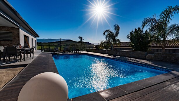 Ein Schwimmbad mit blauem Himmel, strahlender Sonne und einer Terrasse mit Gartenmöbeln.