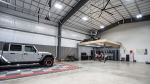 A spacious indoor garage with a large vehicle and a motorcycle, metal beams on the ceiling, and a checkered floor pattern. - Teton Basecamp Barndominium (Driggs)