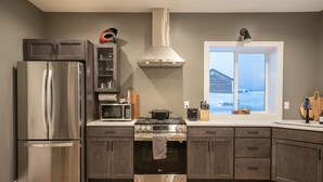 A modern kitchen with stainless steel appliances, a large window, and a view of a wooden house. - Teton Basecamp Barndominium (Driggs)