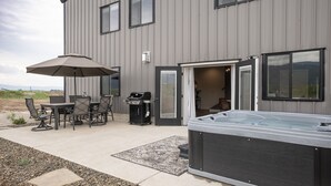 A modern building with a patio area featuring a hot tub, outdoor seating, and a grill. - Teton Basecamp Barndominium (Driggs)