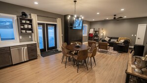 A modern kitchen with a dishwasher, a dining area with a round table and chairs, a living room with a sofa and a TV, and a fireplace. - Teton Basecamp Barndominium (Driggs)