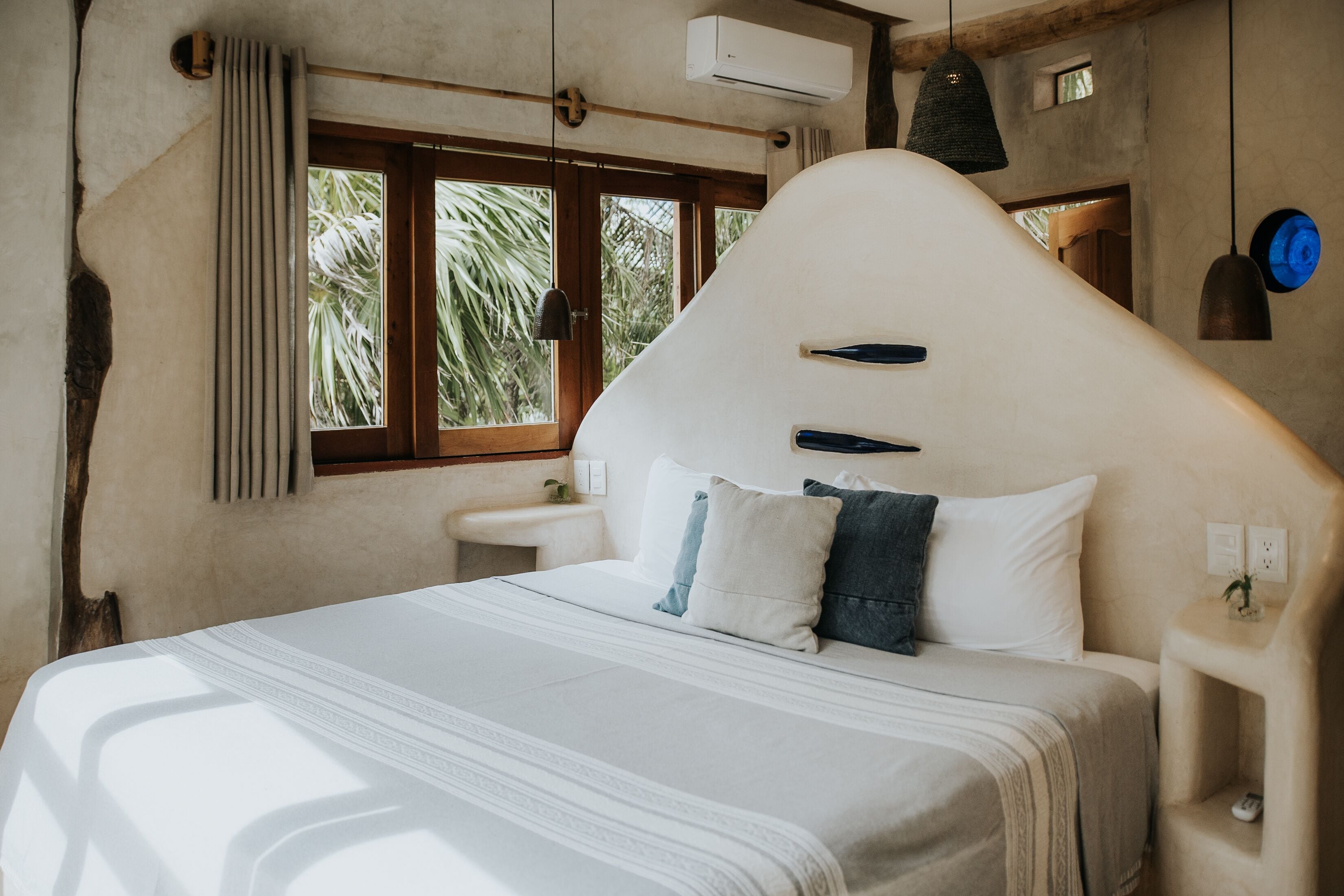 A bedroom with a unique curved headboard, a large window with a view of palm trees, and a bedside table with a lamp.