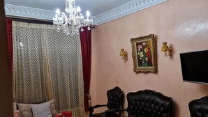 Un salon comprenant un canapé, deux fauteuils, une table basse agrémentée d’objets décoratifs, un tapis rouge à motifs, un lustre et un tableau accroché au mur.