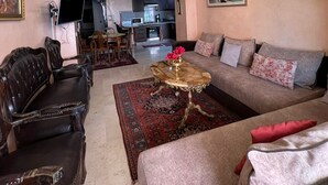 Un salon avec un canapé, des fauteuils, une table basse et un lustre. On aperçoit une cuisine au fond.