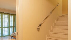 Una escalera con barandilla, suelo de madera y una puerta que da a una habitación con cama y ventanas amplias.