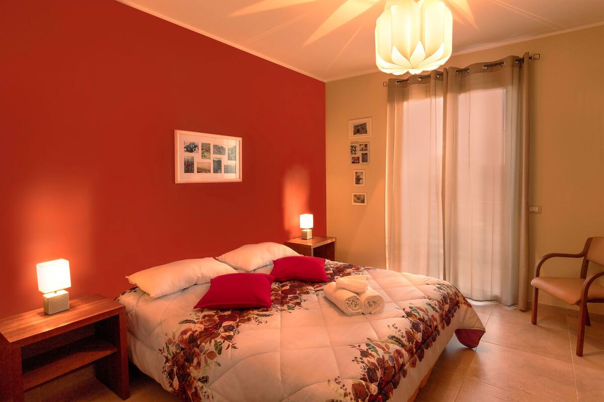 Una cama bien hecha con cubrecama floral, dos almohadas rojas y toallas enrolladas sobre una mesita de noche de madera.