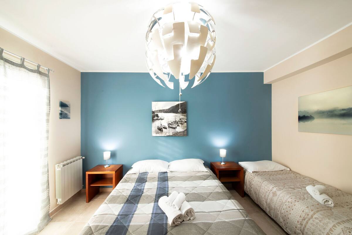 Habitación de hotel con cama, dos mesitas de noche, radiador, lámpara colgante y un cuadro en la pared.
