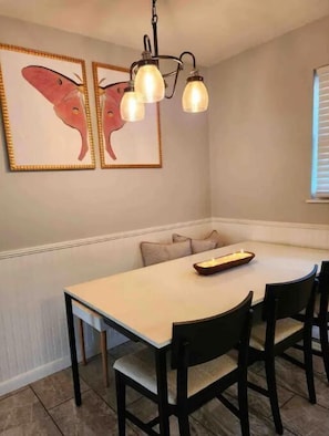 Un comedor con mesa blanca, sillas negras, una araña y dos obras de arte con mariposas enmarcadas en la pared.