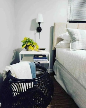 Una habitación de hotel ordenada con una cama, una mesita de noche con una lámpara, un estante pequeño con libros y una cesta tejida negra.