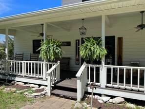 Un porche blanco con barandilla, dos macetas, un banco y una puerta.