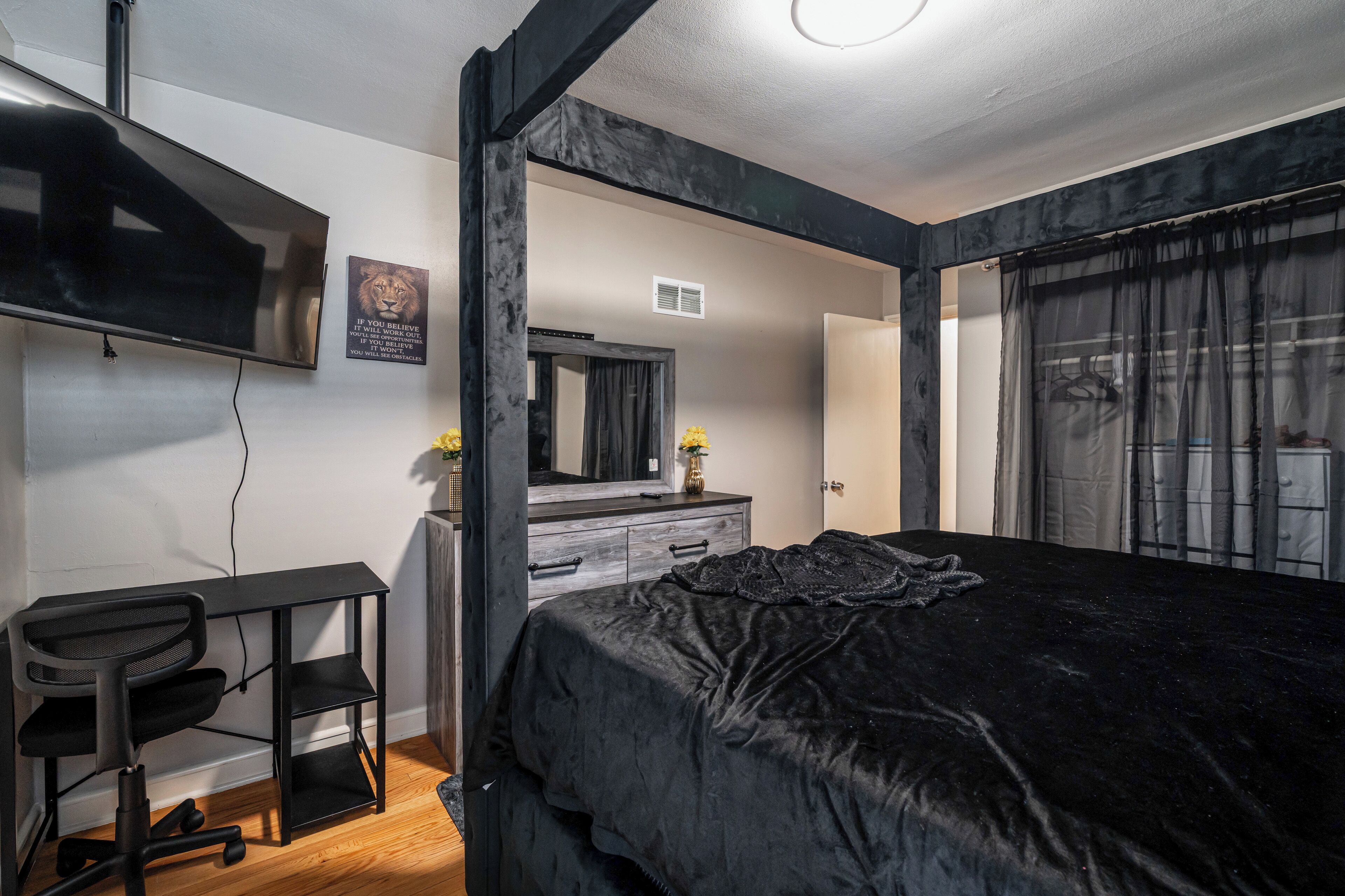 Une chambre à coucher comprenant un lit, un bureau, une chaise, une télévision, une commode, un miroir et une fenêtre avec des rideaux.