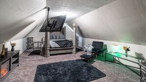 Una camera da letto con soffitto spiovente, un letto con biancheria grigia, un tappeto nero, un tavolino in vetro e una sedia.