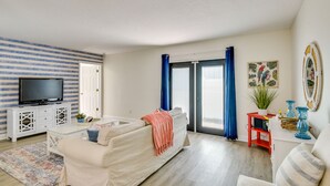 A living room with a sofa, a TV on a white cabinet, a red side table with a plant, and a large window with blue curtains. - New Smyrna Beach Vacation Rental < 1/2 Mi to Beach (New Smyrna Beach)