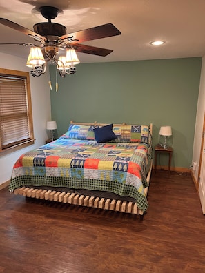 Ein Schlafzimmer mit einem hölzernen Bett, einer bunten Steudecke, einem Deckenventilator und einem Fenster mit Jalousien.
