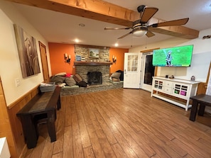 Ein Wohnzimmer mit Kamin aus Stein, Holzboden, Deckenventilator, Fernseher und einer Bank.