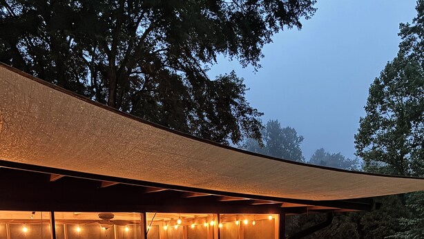 A covered outdoor deck with seating and a table, surrounded by trees. - Mountain Lake House - Hot tub, fire pit, fish and swim! (Almond)